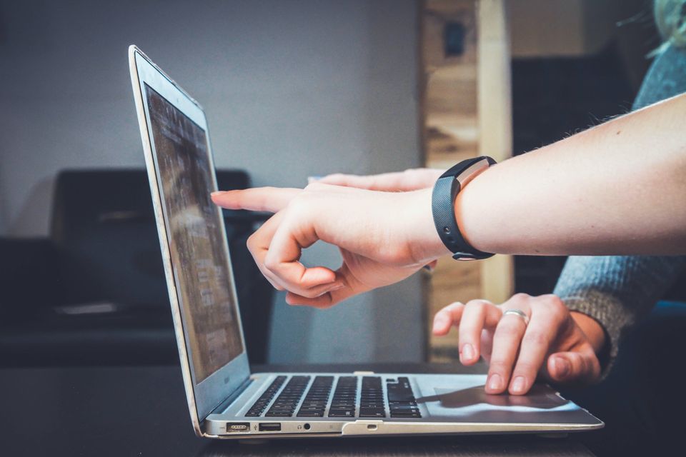 A laptop rests on a table, where one person's hand if pointing to the screen, and another person uses the trackpad.